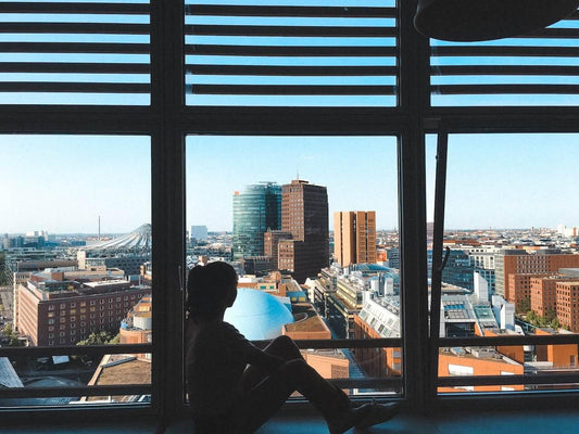 a sad and lonely woman who suffers from social anxiety disorder sitting on the floor in her apartment looking at the buildings across the street through the window